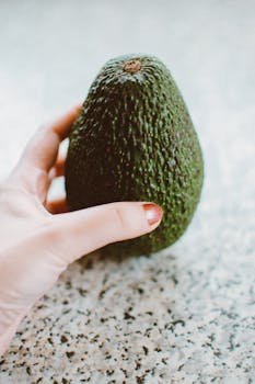 A hand with painted nails holding a ripe Hass avocado on a speckled surface for a healthy lifestyle concept.